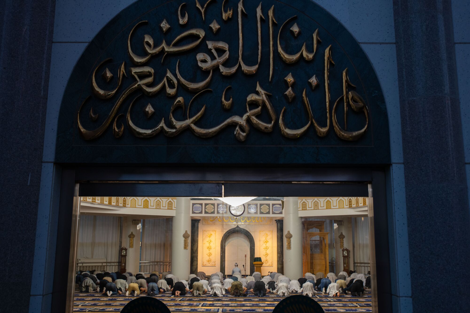 A view of people kneeling in prayer beyond an entryway, above which hangs a sign in blue with golden script in Arabic 