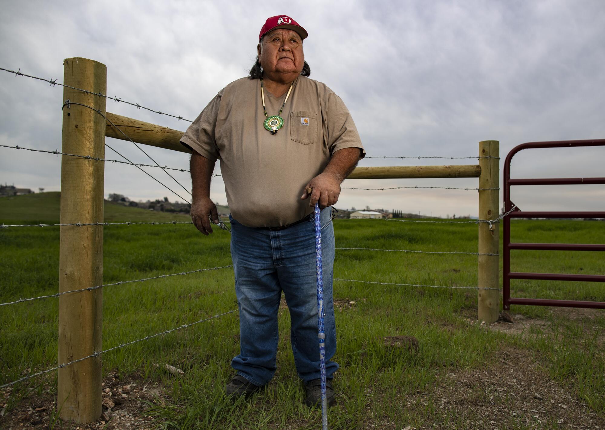 A man in a beaded pendant and red baseball cap, holding a cane and standing by a gate and barbed-wire fence in a grassy field