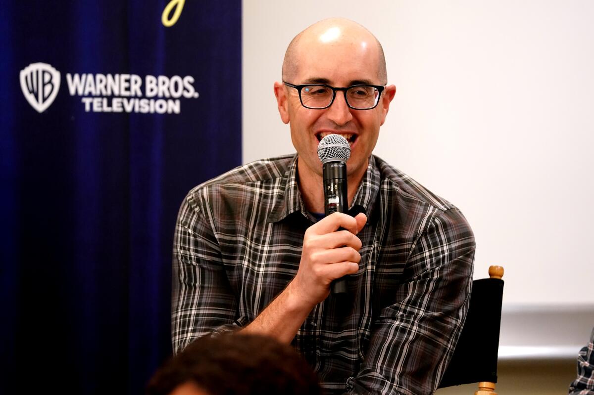 Justin Halpern, wearing black-rimmed glasses and a plaid brown shirt, holds a microphone to his mouth.