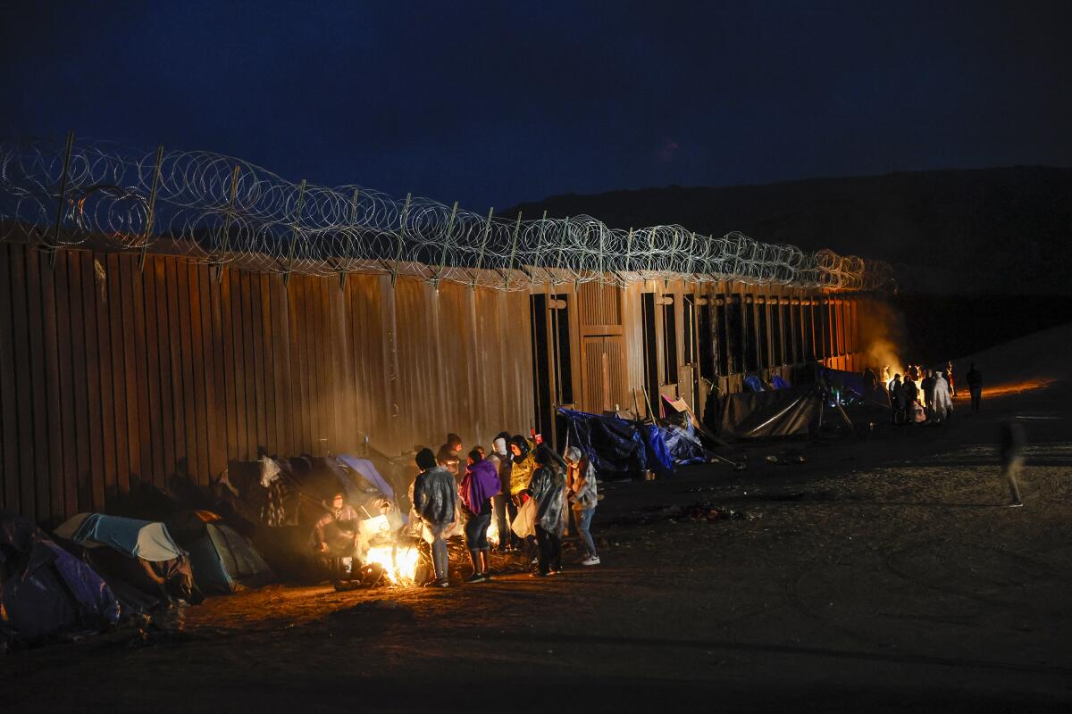 People gather by a fire in front of a wall topped by barbed wire