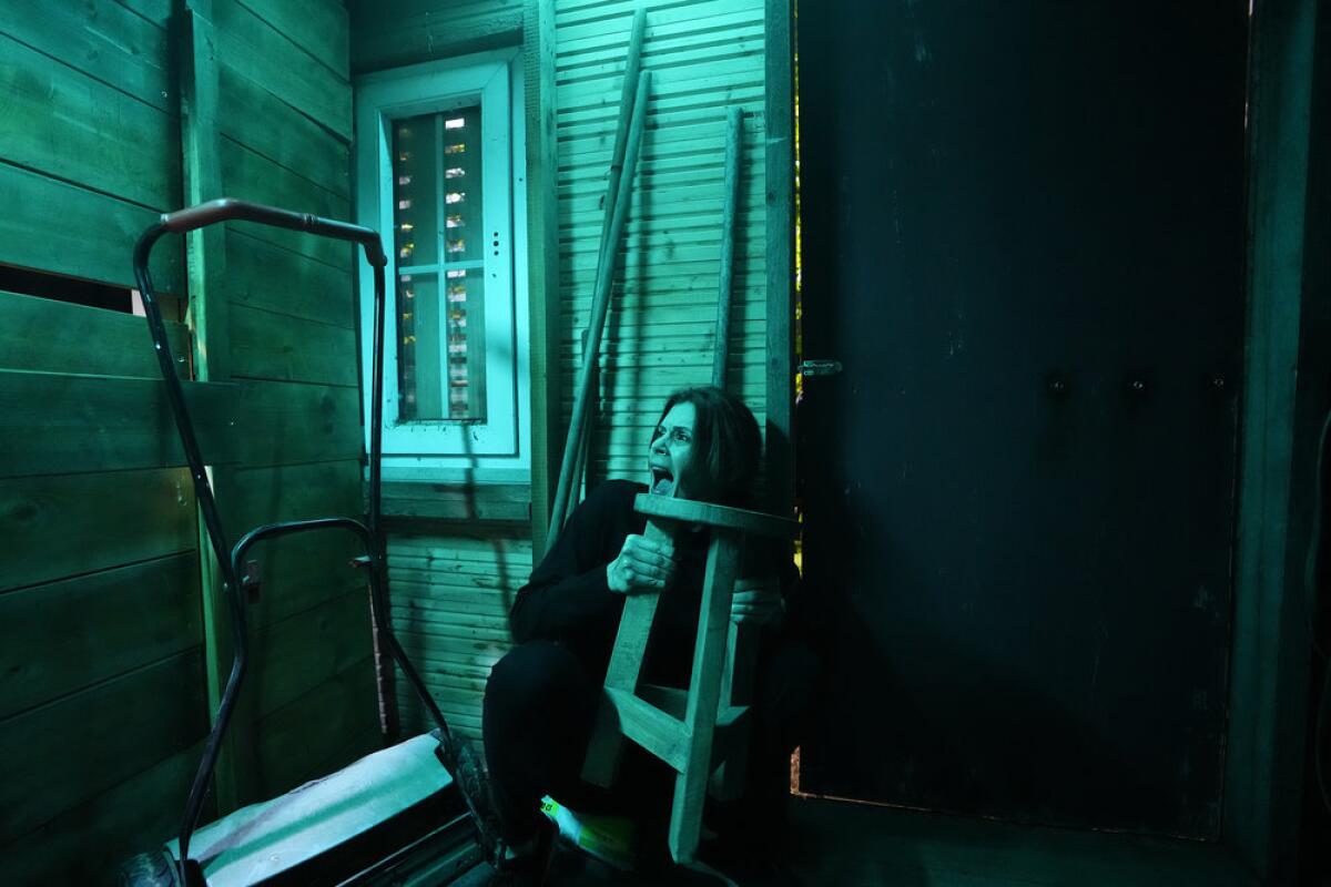 A woman crouches behind a stool in a room lit with blue-green light.