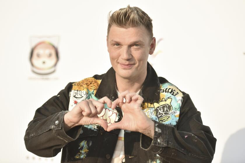 A man with short blond hair wearing a jean jacket and forming a heart shape with his hands