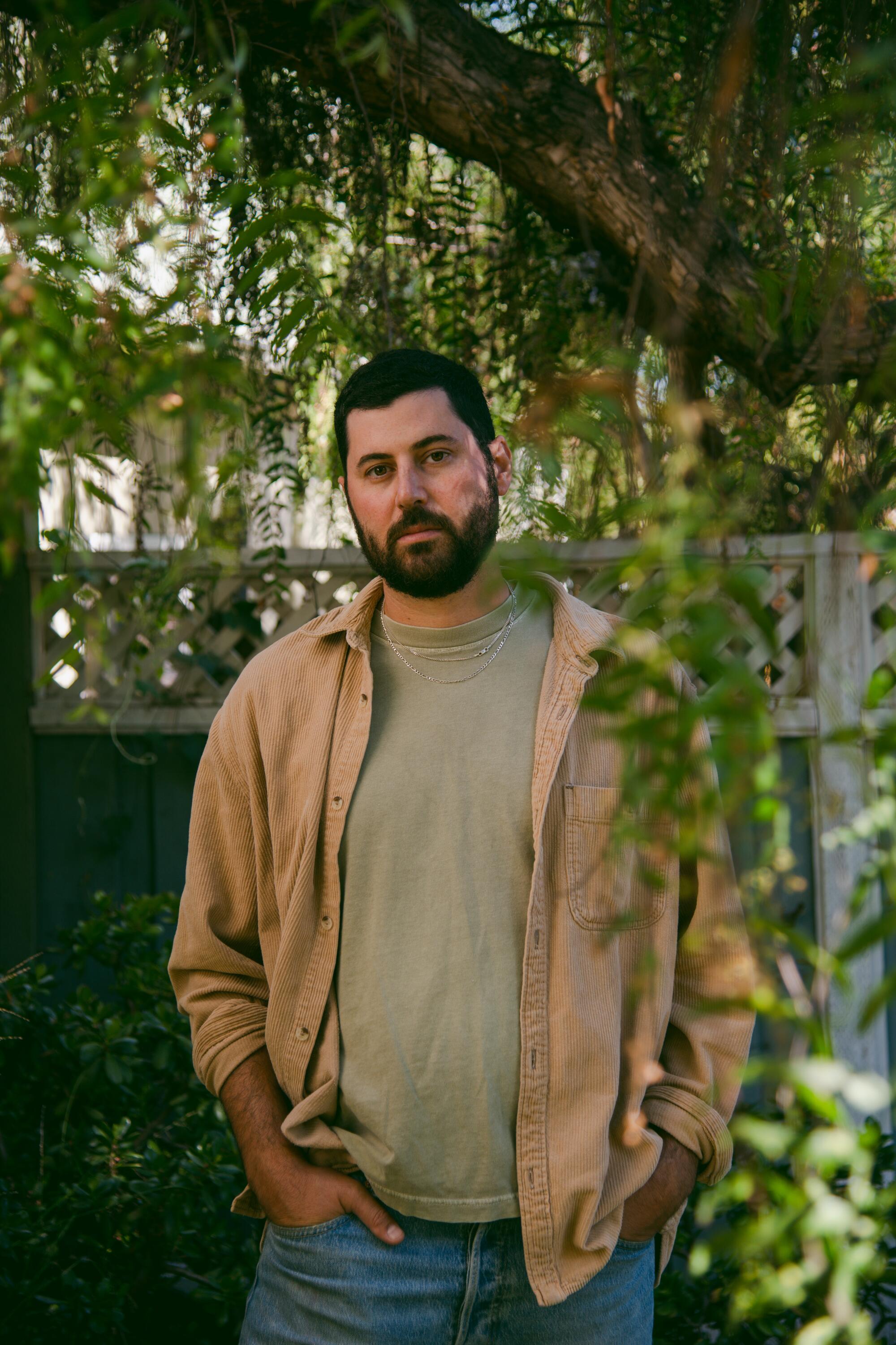 A bearded man in a gray tee shirt, tan jacket and jeans standing in a garden with an overhanging tree.