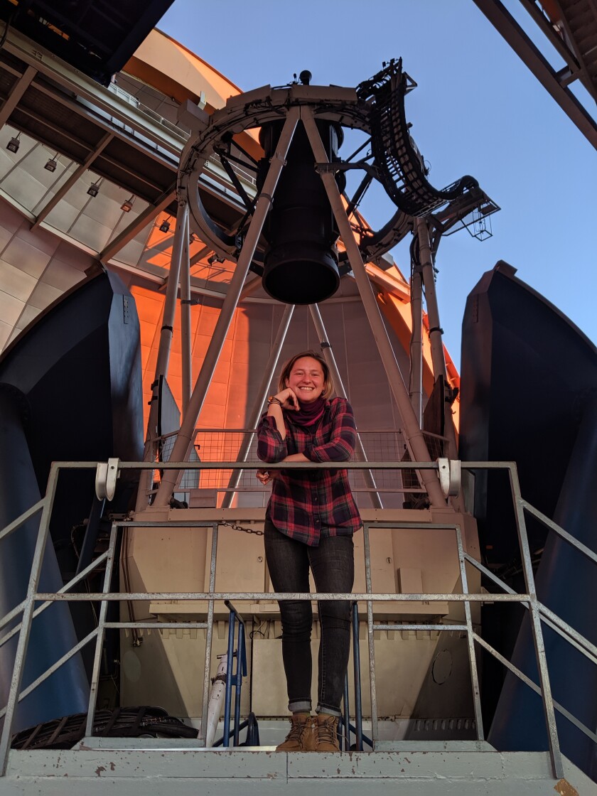 Aliza Beveridge stands in front of a large telescope in an open roof observatory.