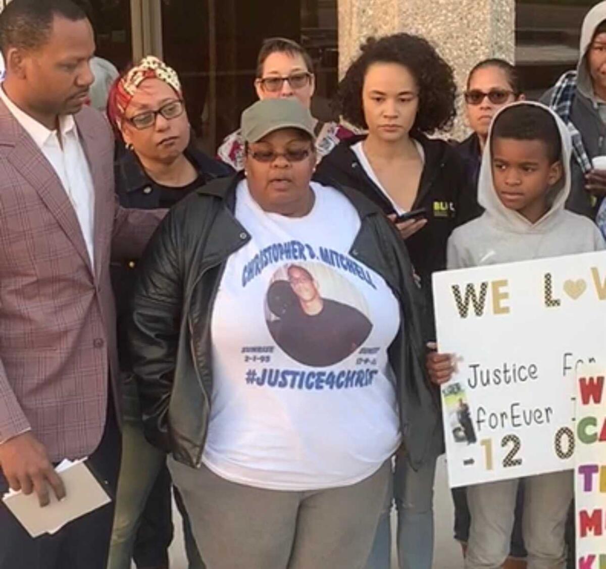 A woman in a white "#Justice4Chris" shirt stands speaking, flanked by several people including a child with a homemade sign
