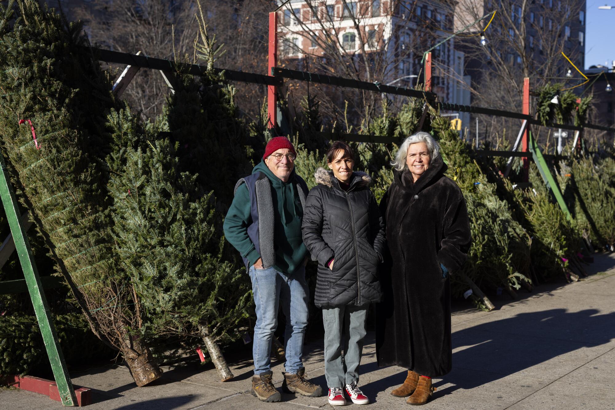 Three people stand in front of a row of cut Christmas trees.