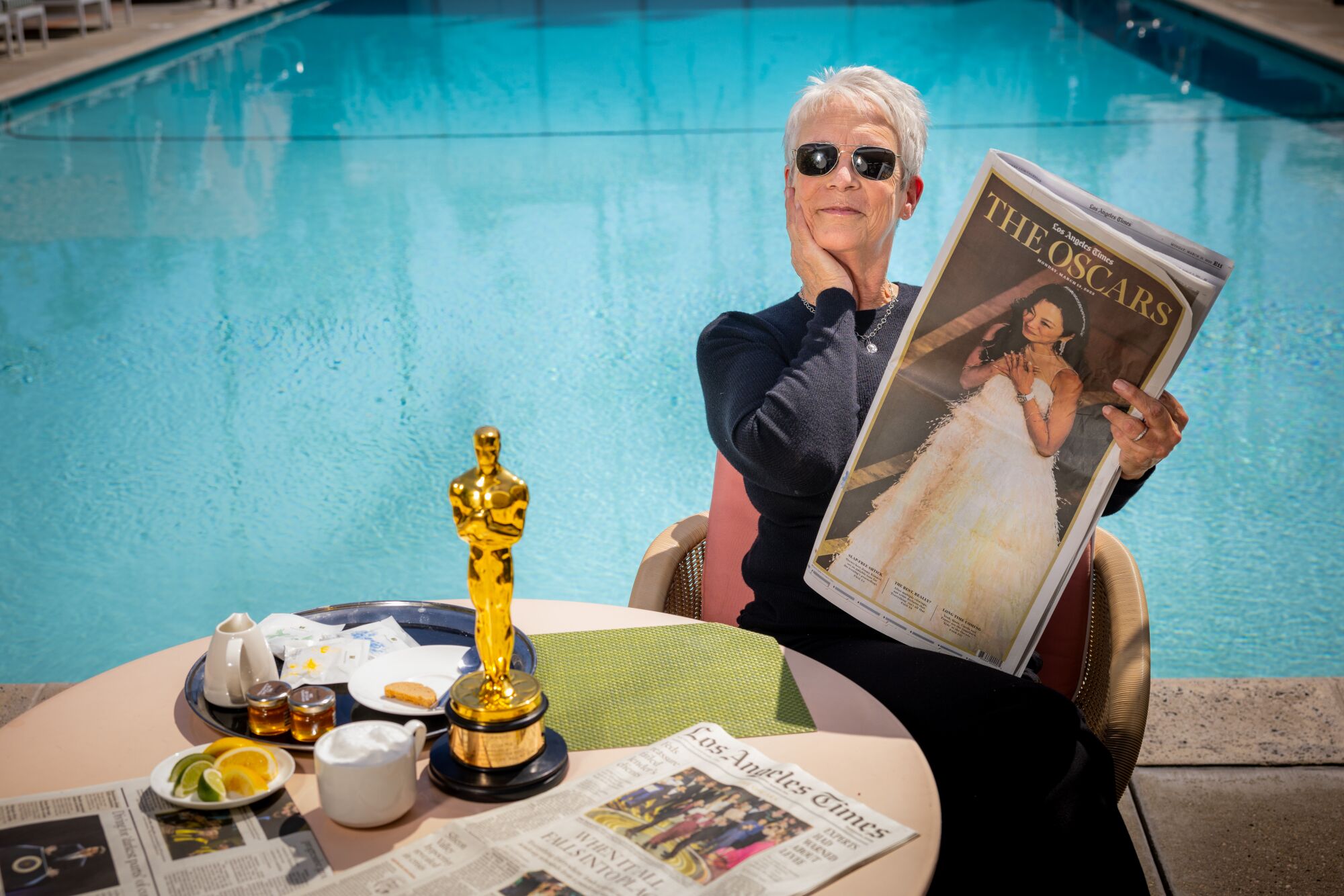 A woman in sunglasses holds a newspaper clipping next to a table on which her Oscar is written.