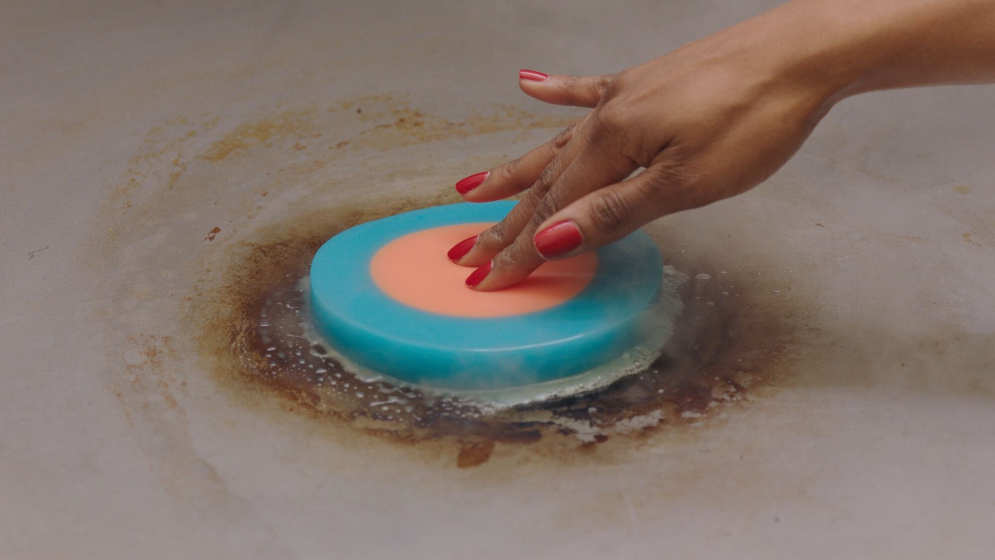 A circular pancake of dense blue and peach gelatin is pushed into a pan by a woman's hand with painted red nails.