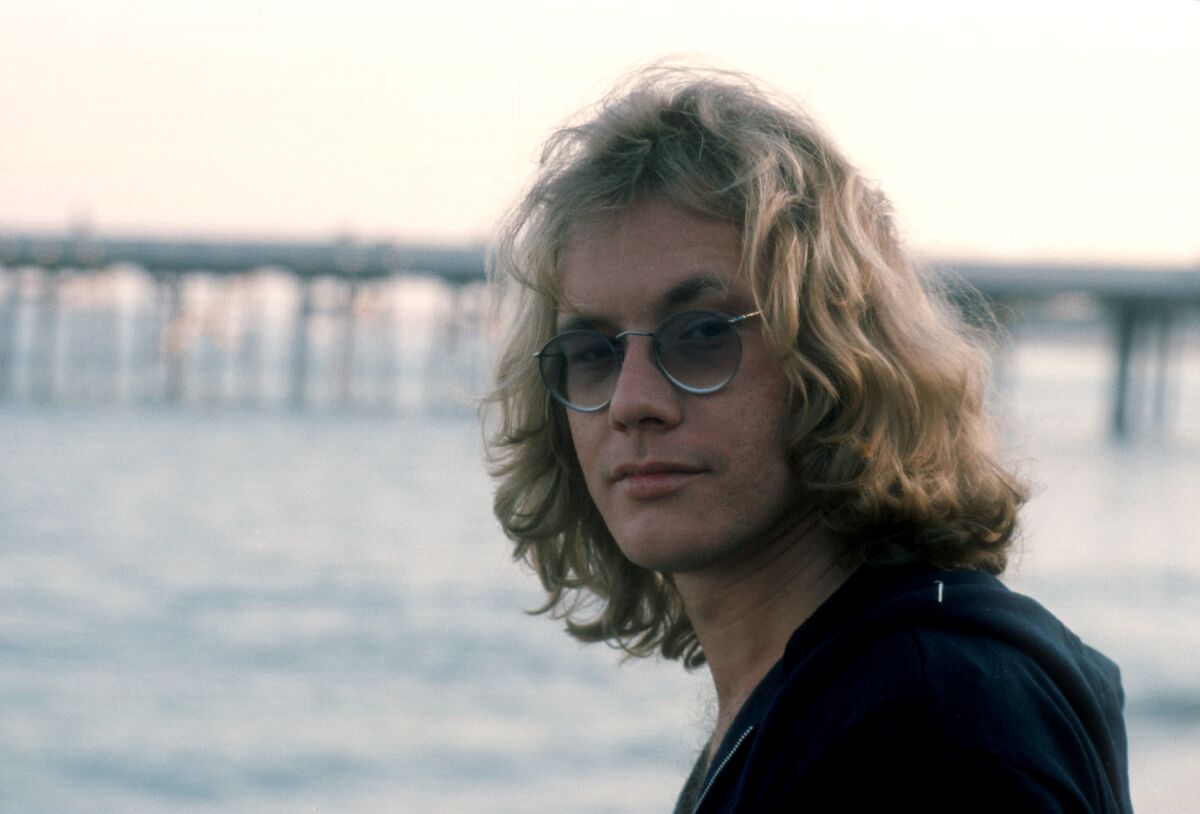 A close-up portrait of a man with sunglasses and shoulder-length blond hair, with a pier and ocean in the background
