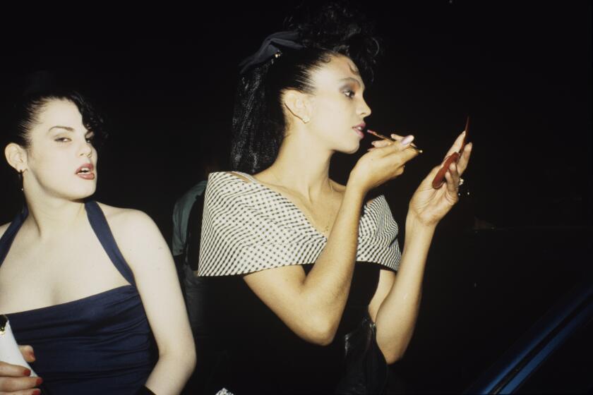 Carmen Xtravaganza stands next to another person, wearing a black outfit, staring into a compact mirror, applying makeup 