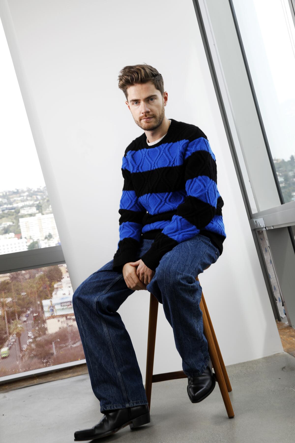 A man wearing a blue striped sweater and jeans sits on a stool.