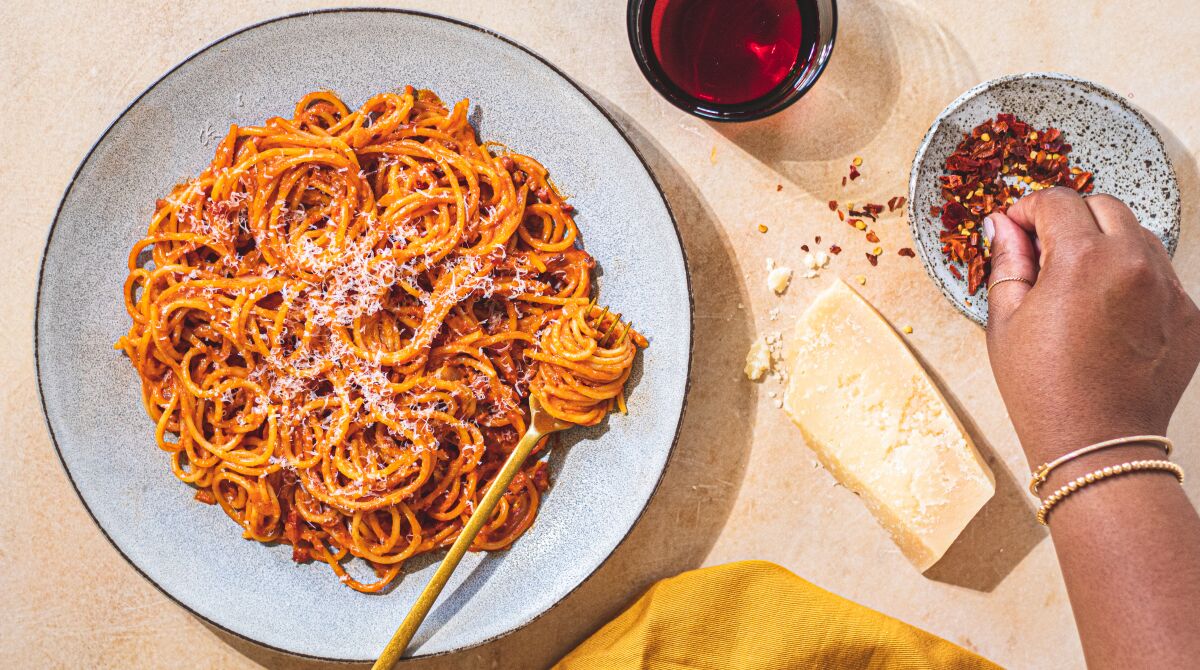 A hand reaches for red pepper flakes to add to a plate of spaghetti in a tomato sauce