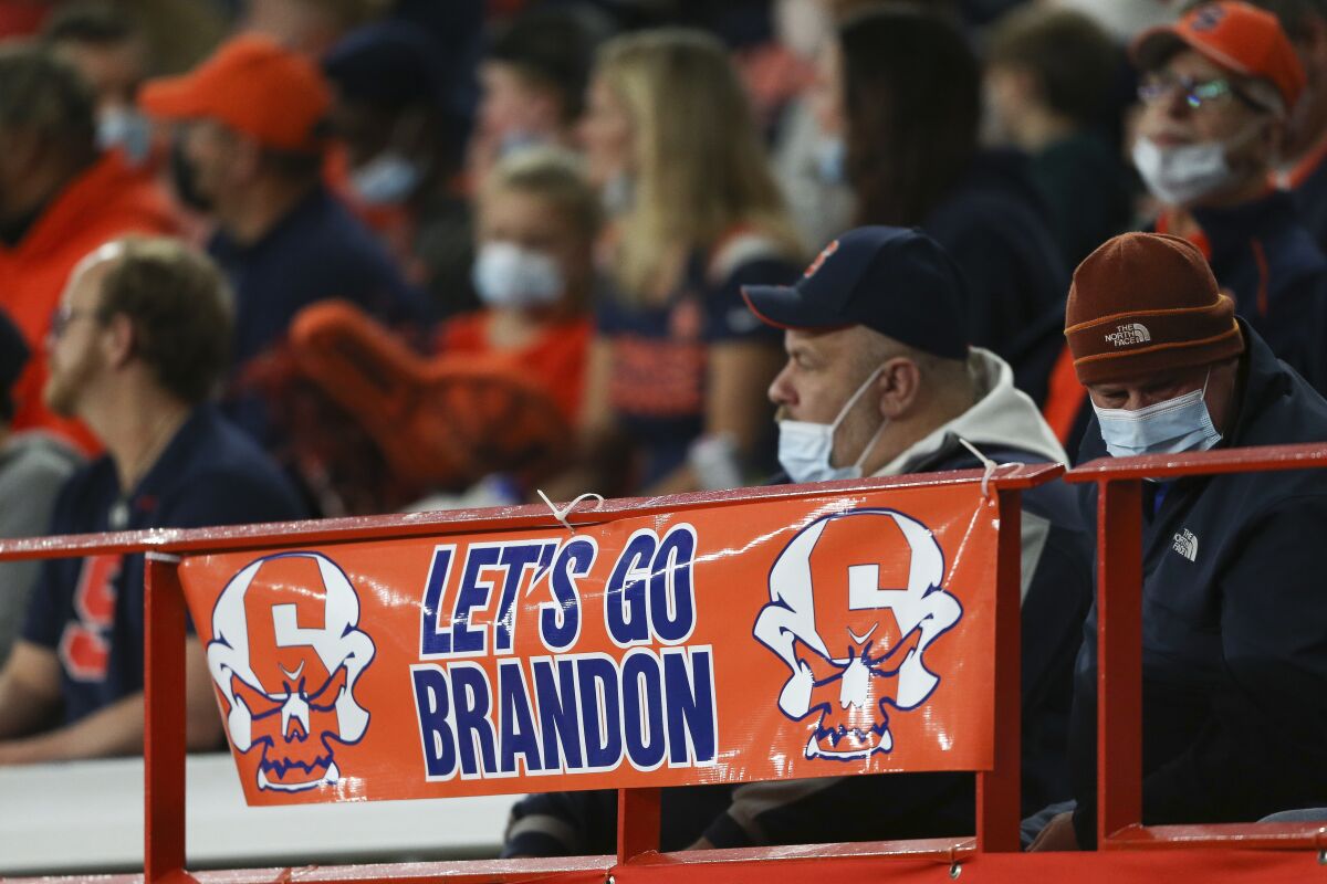 A man with a mask below his nose sits behind a banner reading "Let's go Brandon" at a college football game.