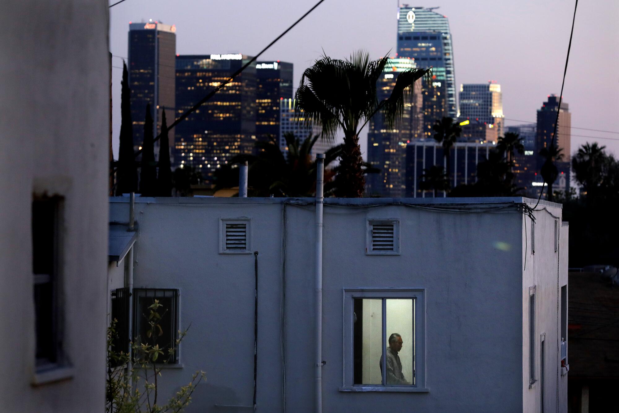 A man is seen in the window of his Los Angeles apartment, with a view of downtown L.A. in the distance. 