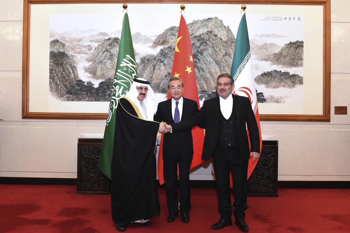 Three men stand side by side and two of them shake hands while three flags stand in the background.