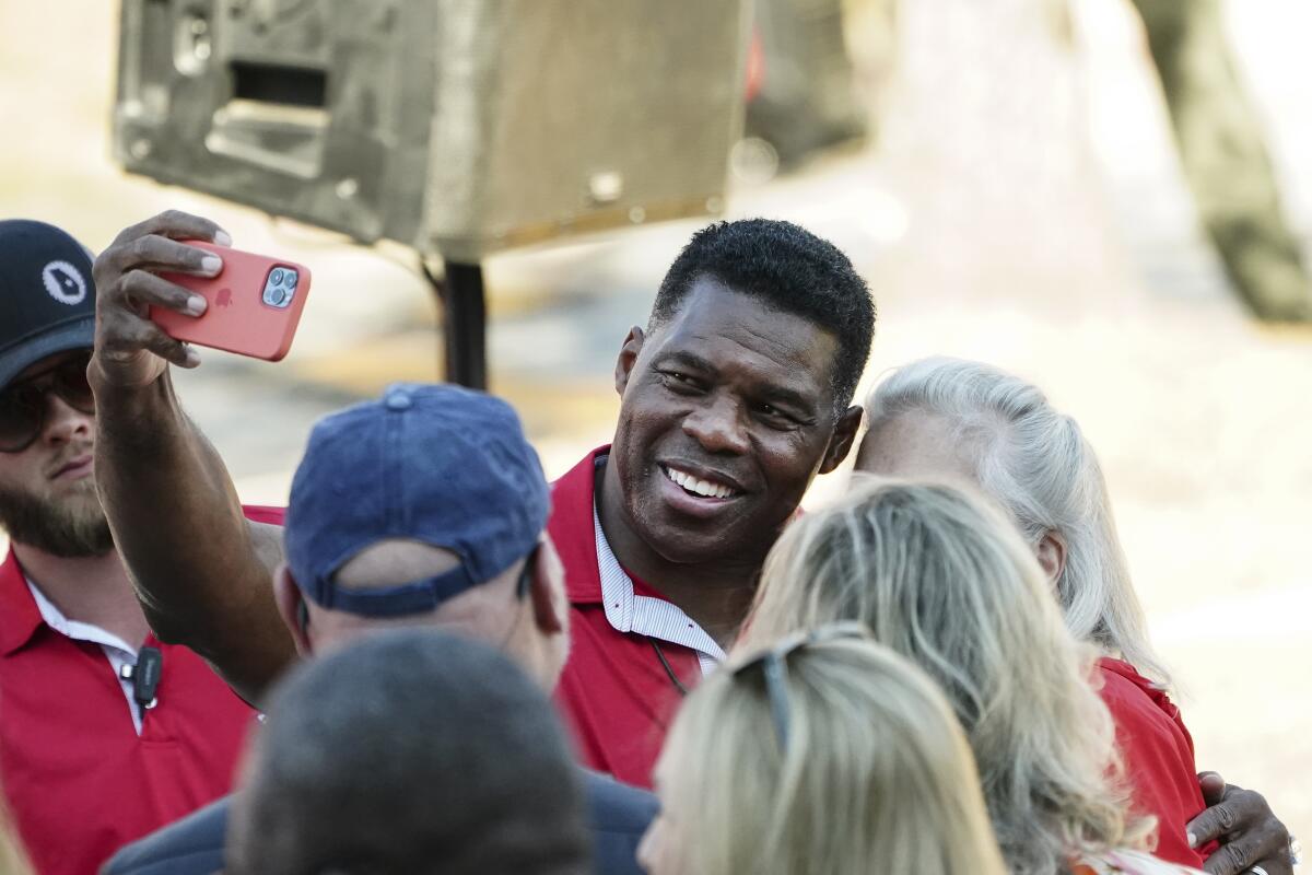 Republican Senate candidate Herschel Walker smiles as he takes a selfie with a supporter as others gather around him. 
