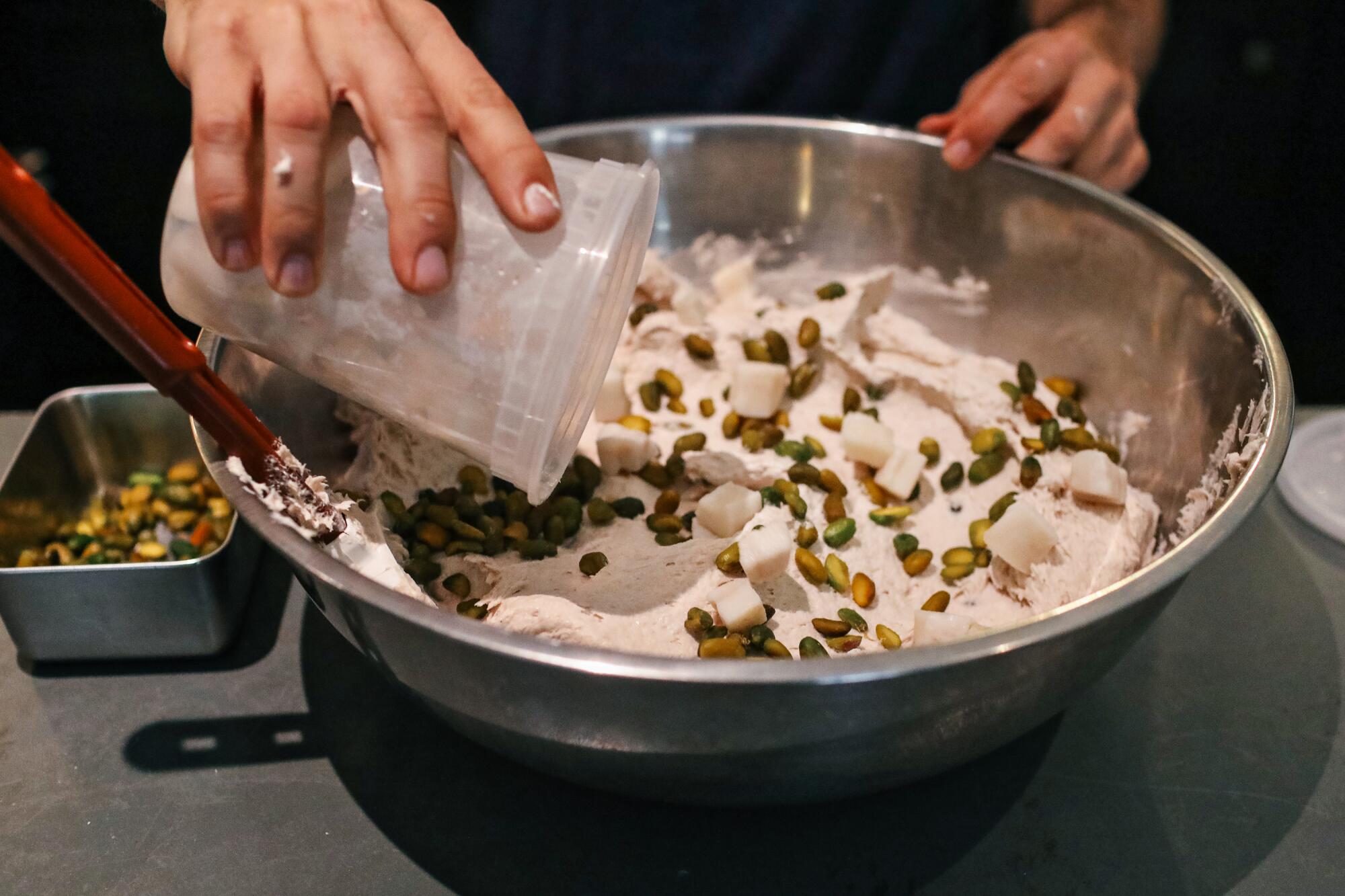 A hand pours a plastic cup of pork fat cubes and pistachios into the emulsified mortadella mix.