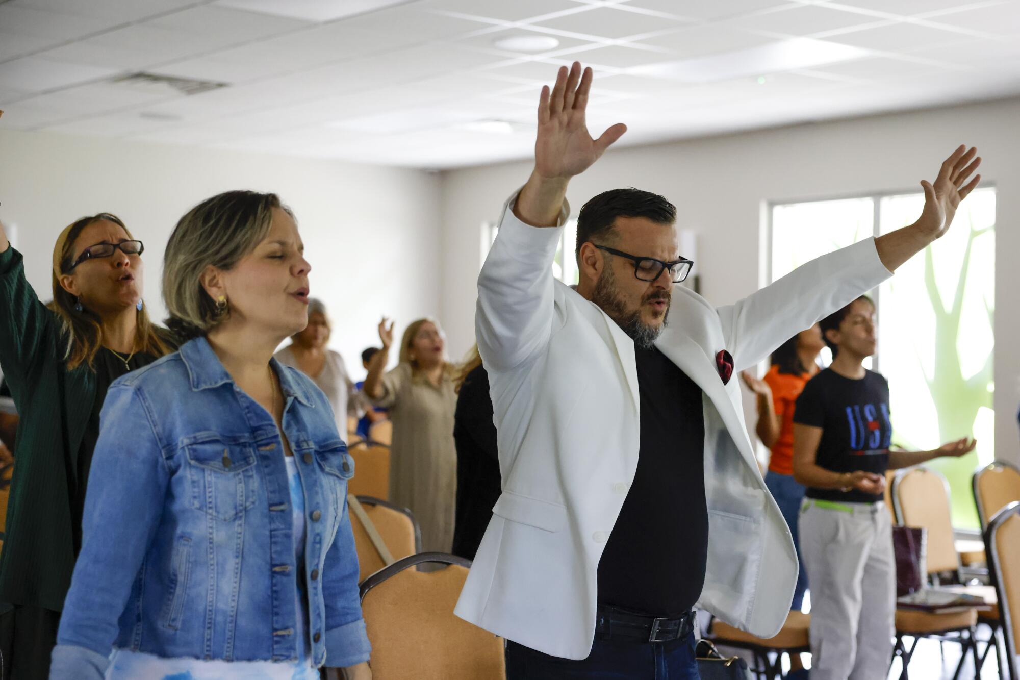 A pastor in a white jacket raises both arms in prayer in a Florida church. 