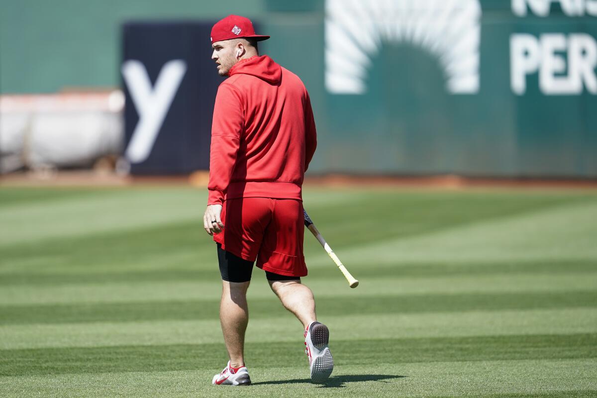 Mike Trout in red shorts and a hoodie walks across a field holding a bat