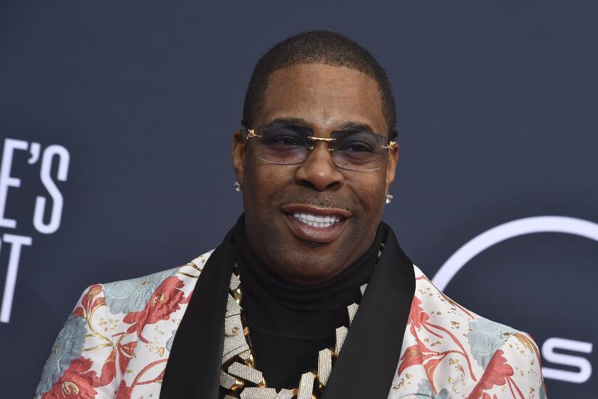 Busta Rhymes in in a pink and blue patterned blazer, chain and sunglasses smiling in front of a dark backdrop