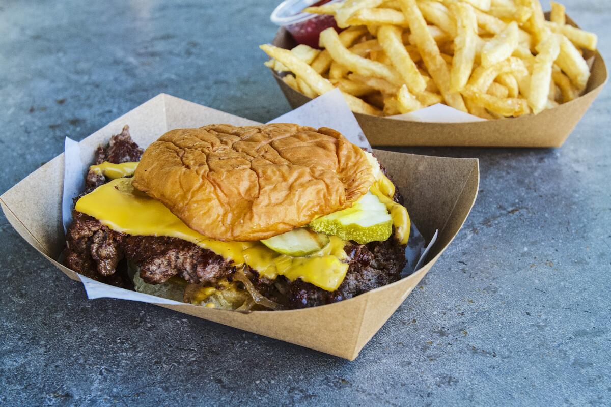 A smashburger made with American Wagyu beef and french fries in side-by-side cardboard dishes.