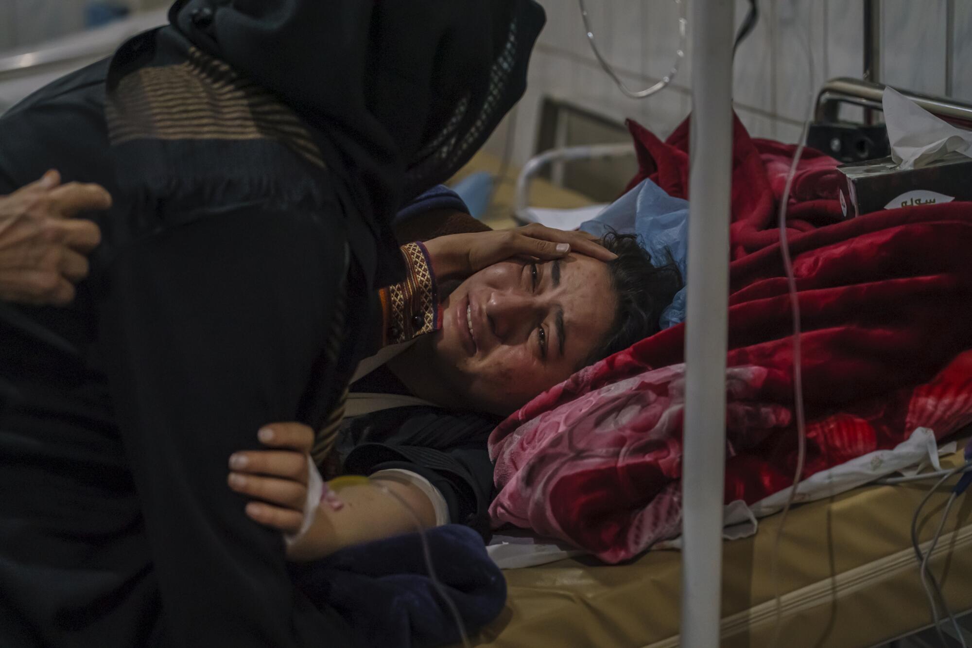 The anguished face of a woman lying in a hospital-type bed is seen as a woman whose head is covered touches her face.