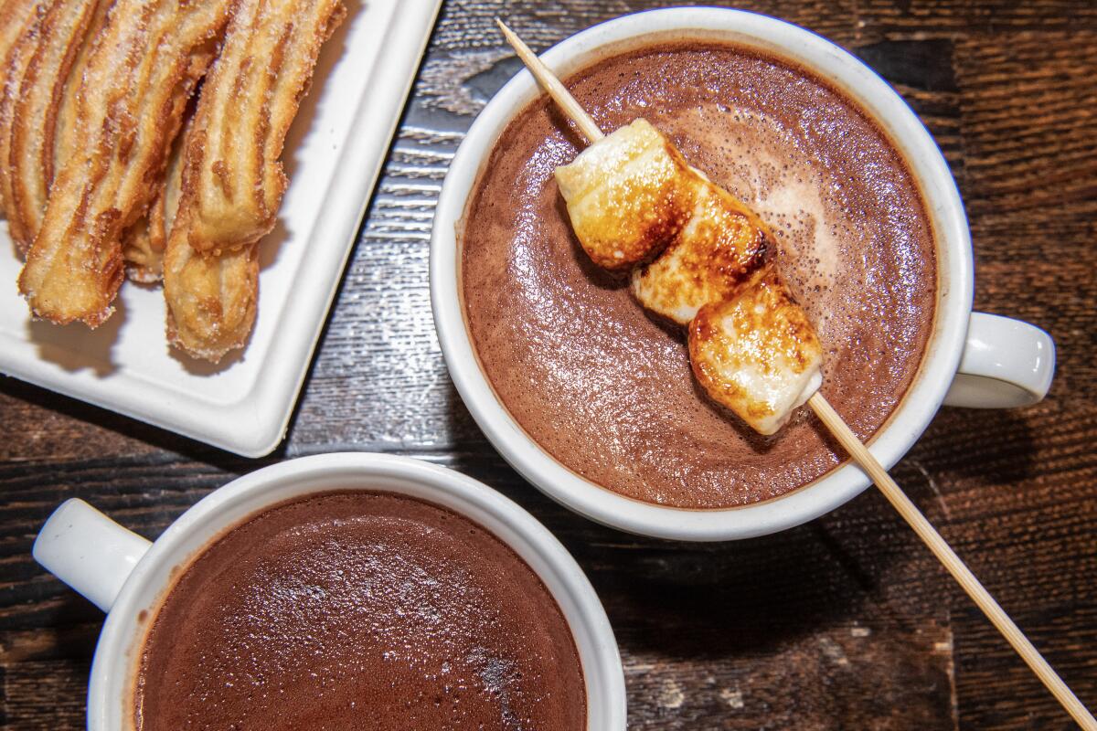 Mugs of drinking chocolate, one with a skewer of toasted marshmallows resting on it, with a plate of churros