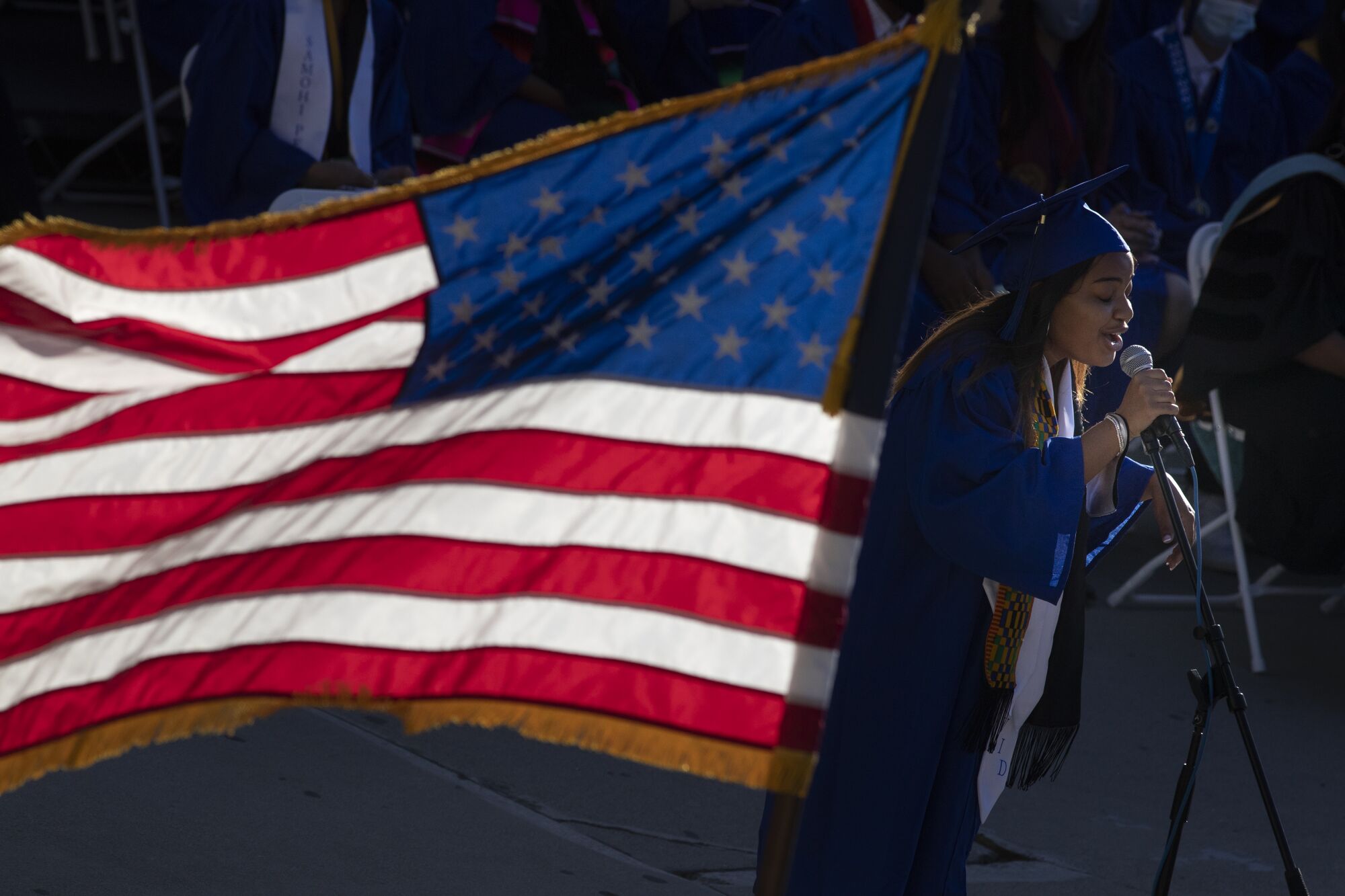 A student in cap and gown sings next to a U.S. flag.