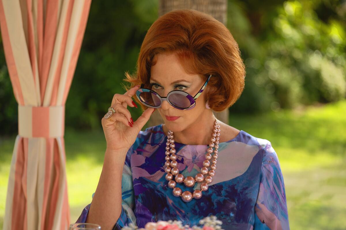 A woman with red hair and a floral top looks over her sunglasses.