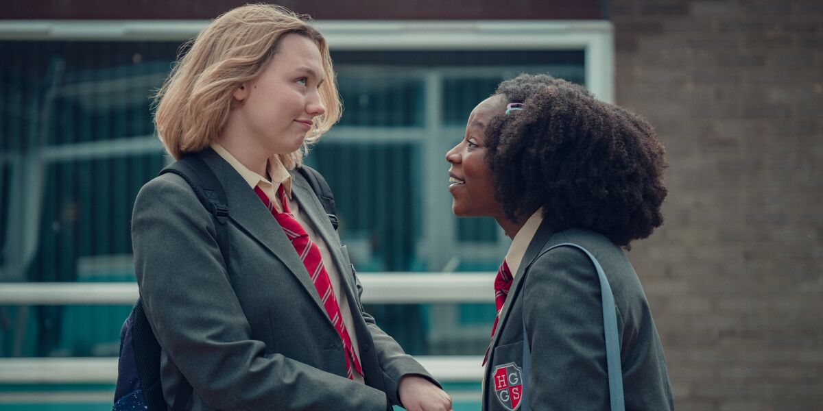 Darcy and Tara, both in school uniforms of gray jackets and red ties, look at one another.