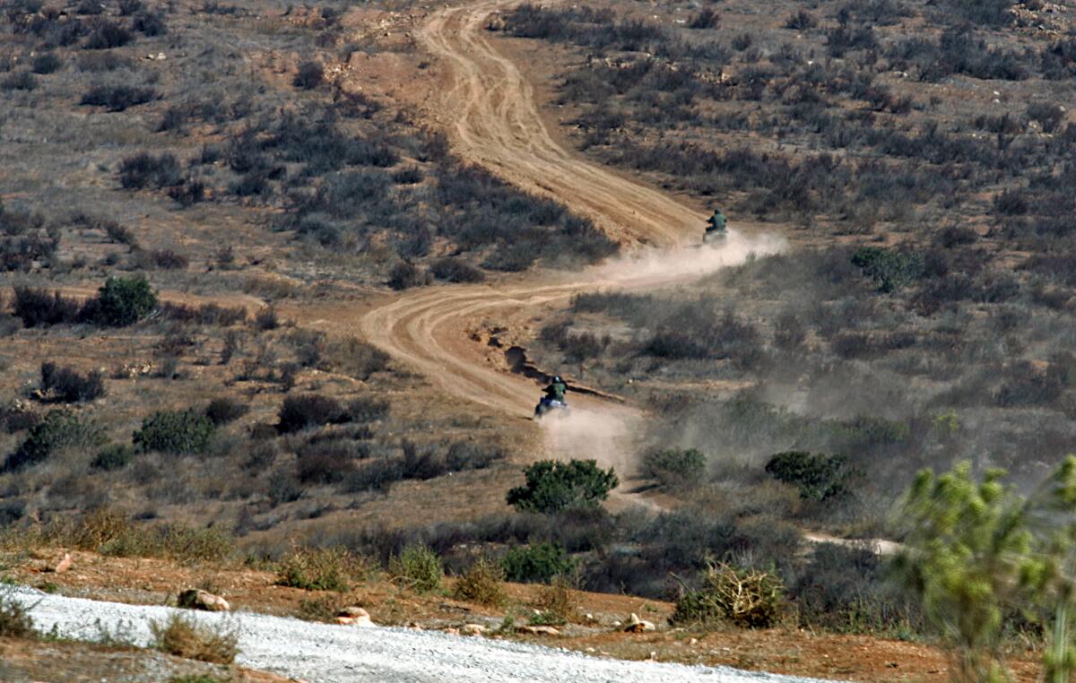 U.S. Border Patrol agents ride ATVs across land filled with shrubs and grass.