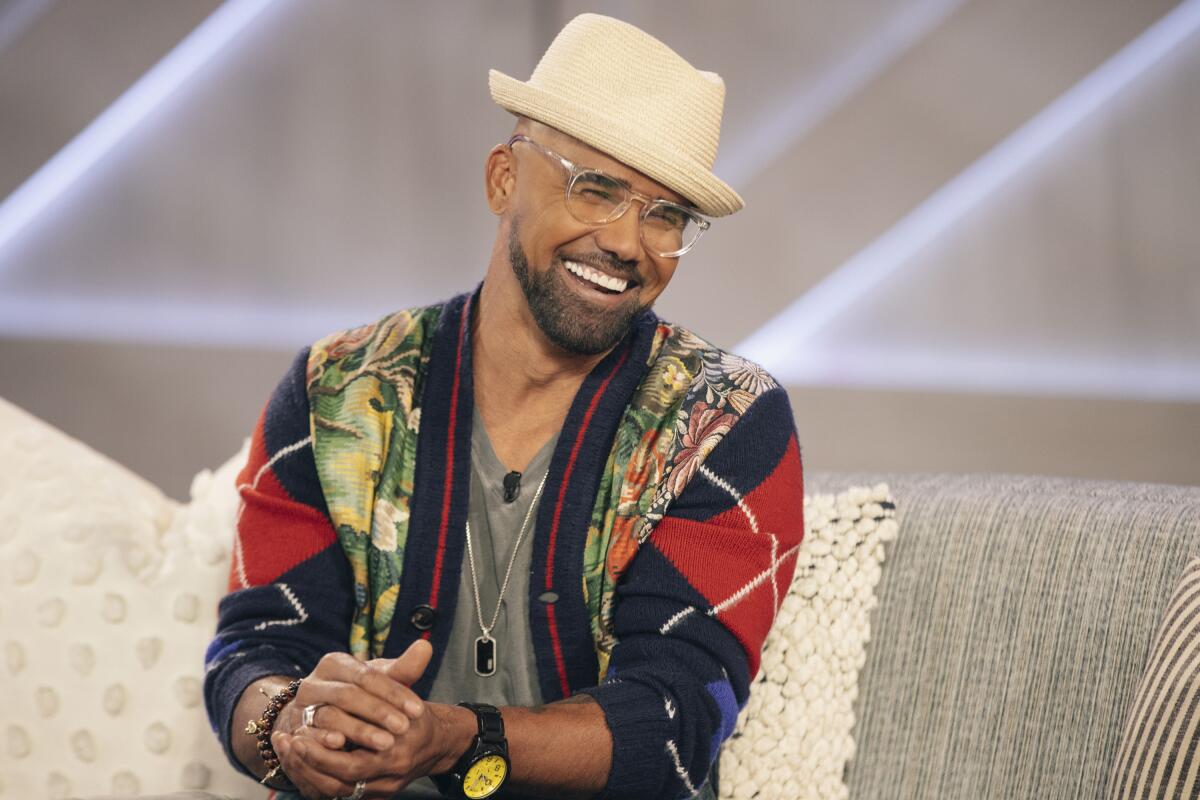 A man with glasses, salt-and-pepper beard and a beige fedora is seated on a couch, smiling.