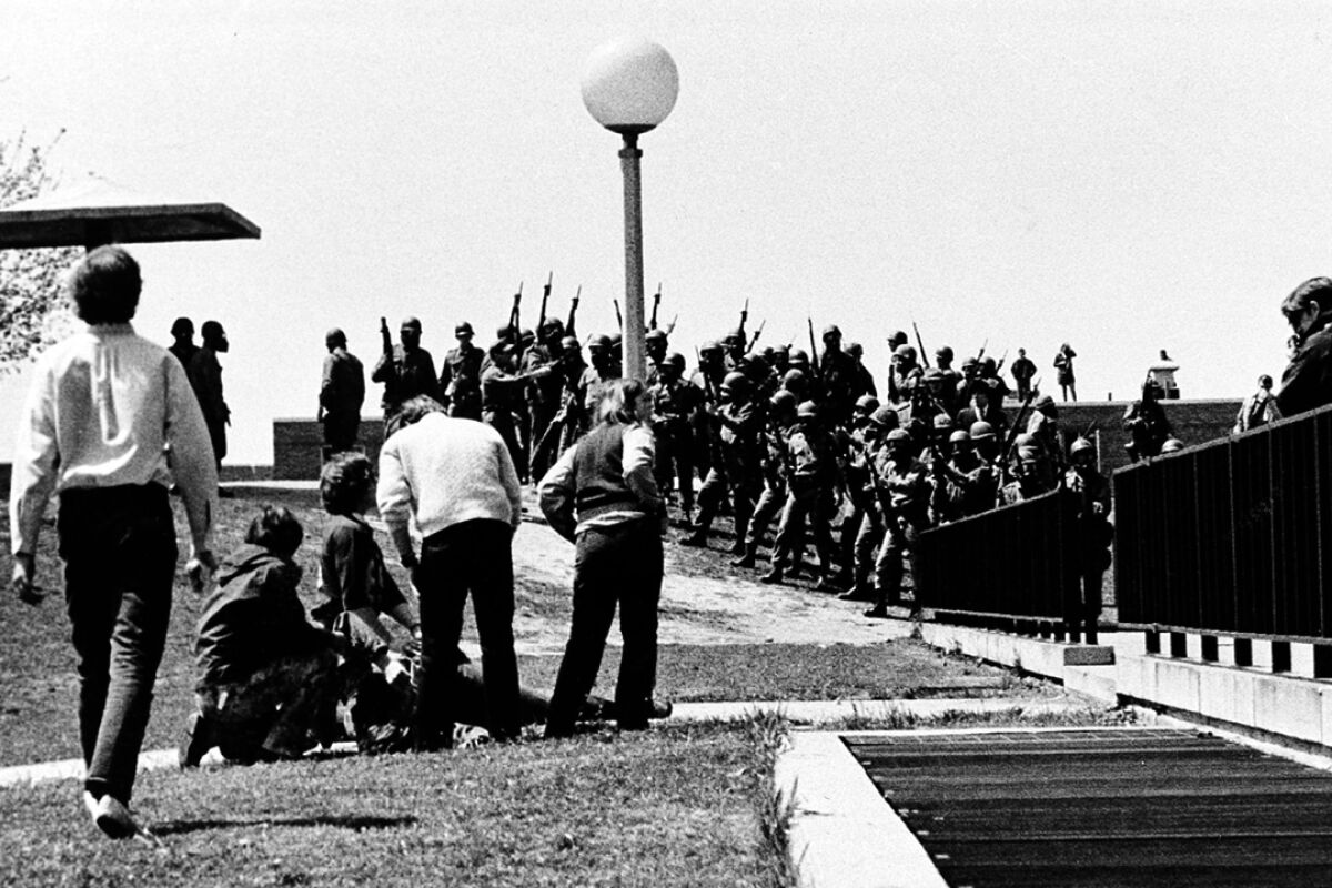 A crowd of men in uniform hold guns in the background. In the foreground, people kneel over a body on the ground.