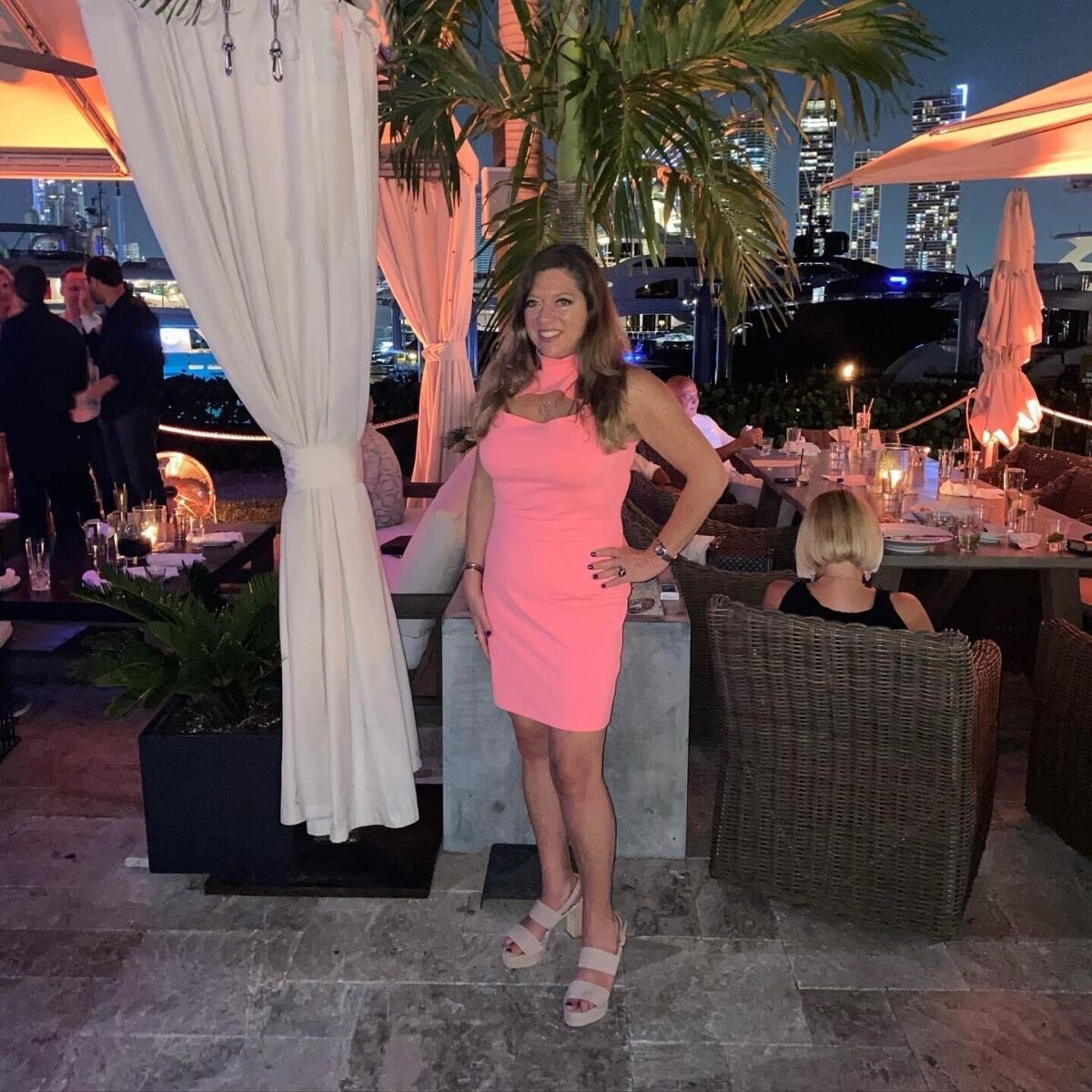 A woman in a coral pink dress stands near diners at tables, with the skyscrapers in the background
