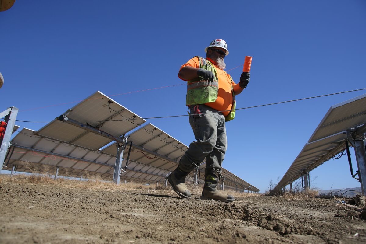 A worker in a vest and hard hat holds a spool near solar panels 