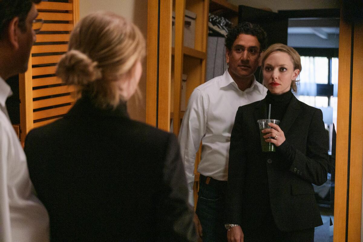 Naveen Andrews in a white button-down and Amanda Seyfried in a black suit look at themselves in a mirror.