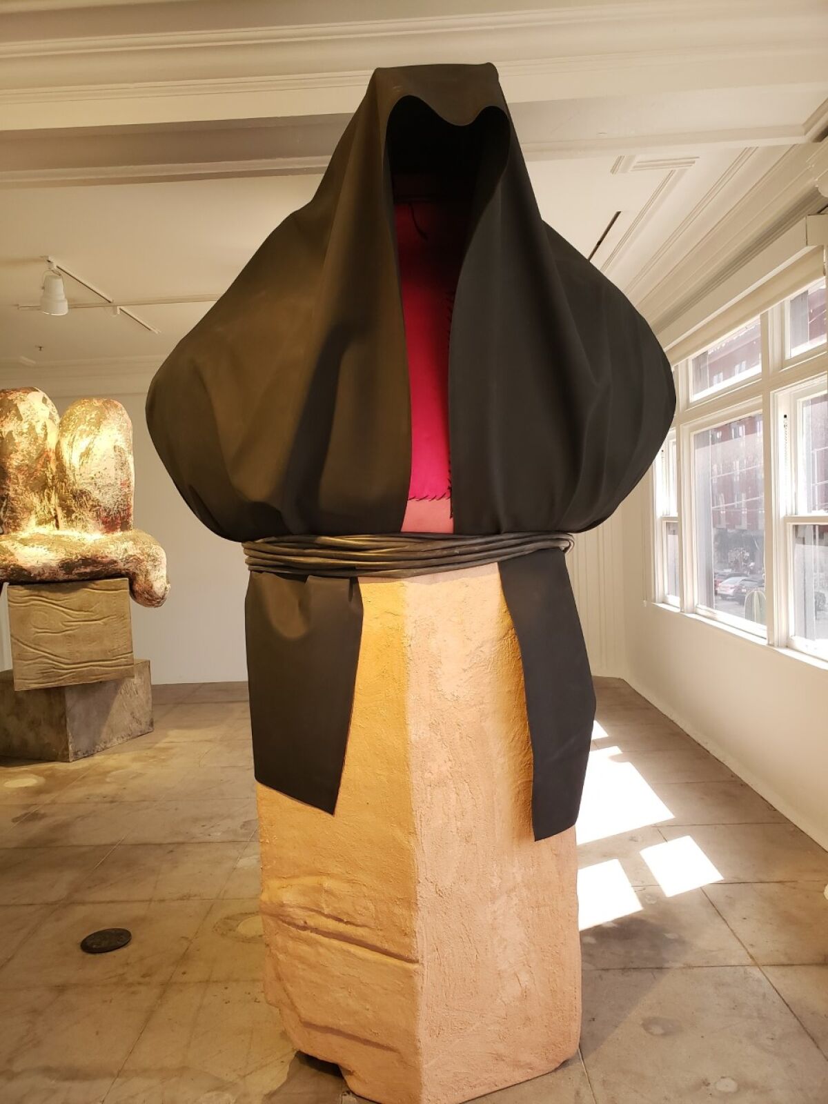 A dark cowl tops a blocky rectangular base. The sculpture sits next to a window.