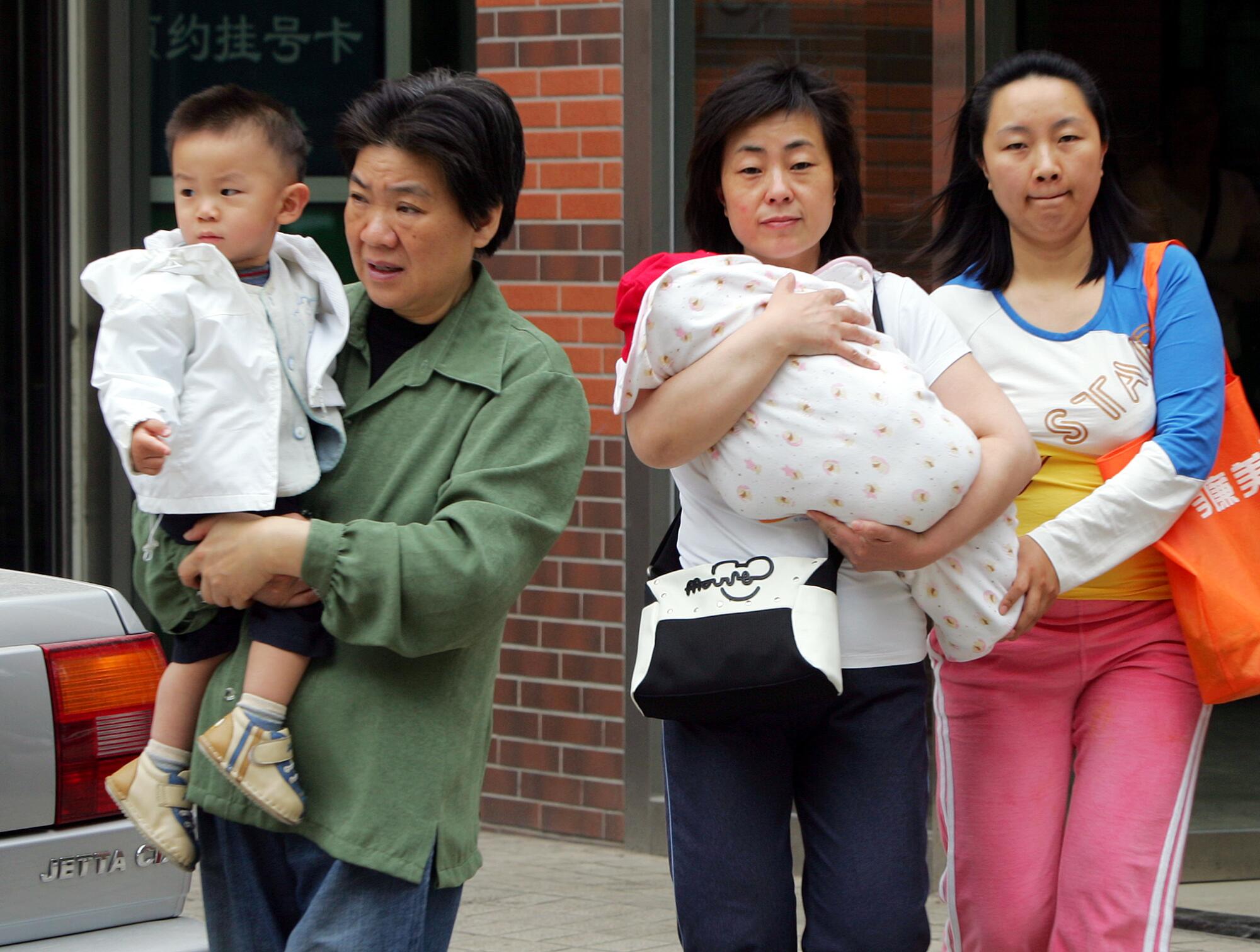 زنی پسری را در سمت چپ نگه می‌دارد در حالی که دیگری نوزاد قنداق شده را در کنار زن جوانی در نزدیکی ساختمان نگه می‌دارد.