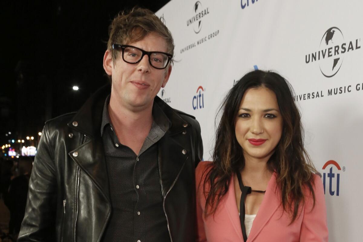 A blond man wearing glasses and a black leather jacket posing with a brunette woman wearing a pink blazer