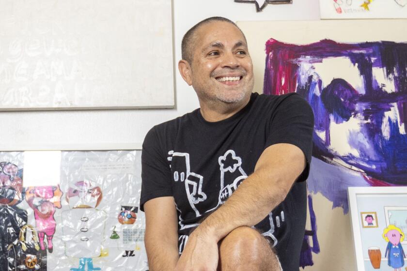 Artist Ricky Sencion in his studio with his artwork behind him and tools for painting in front of him on Dec. 22, 2023 in the Mid-Wilshire area of Los Angeles, Calif. The piece on the top right is called "God, Will I Still Be Gay in Heaven?"