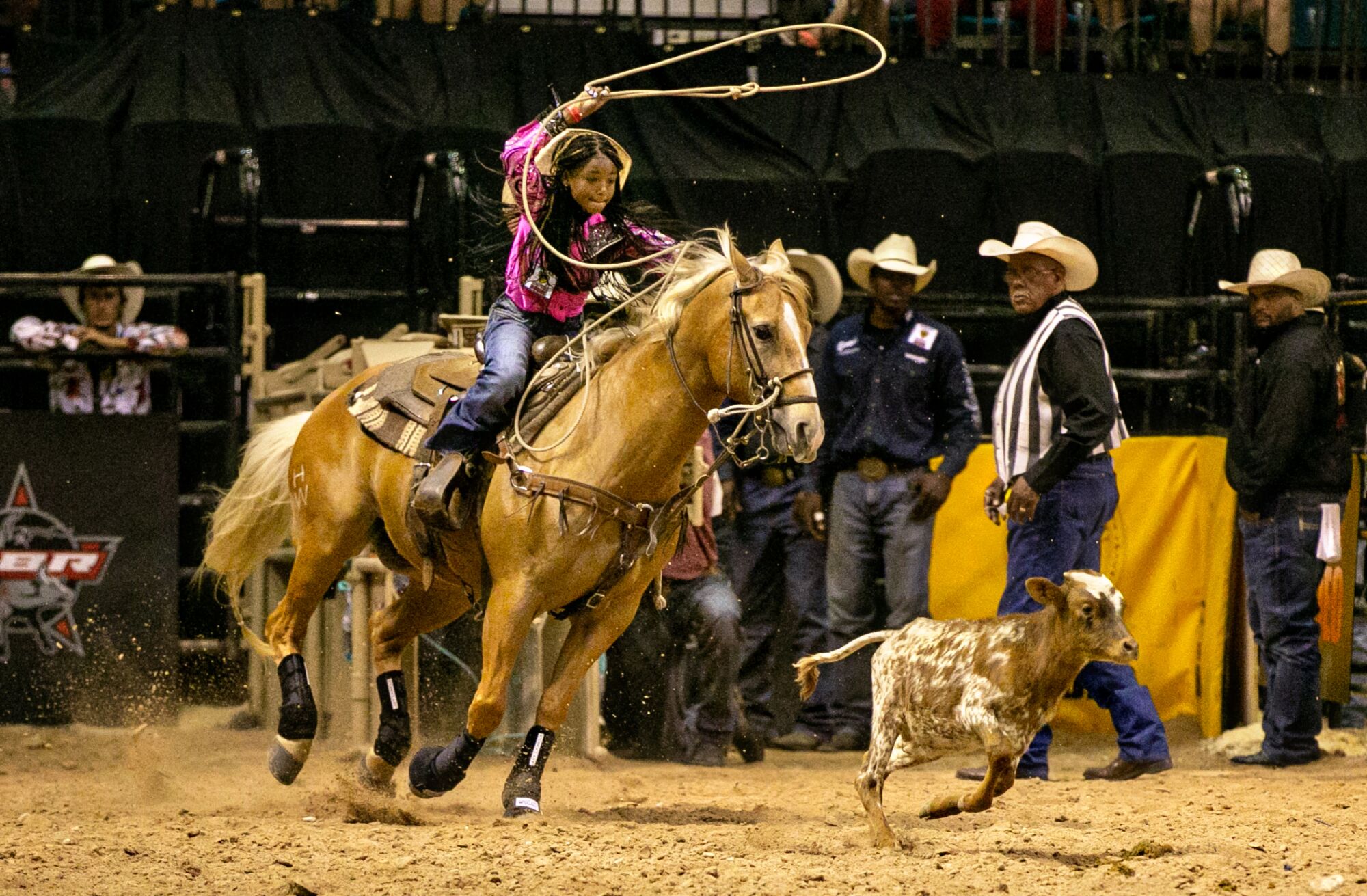 A girl on a horse attempts to lasso a running calf as adults in cowboy hats look on.