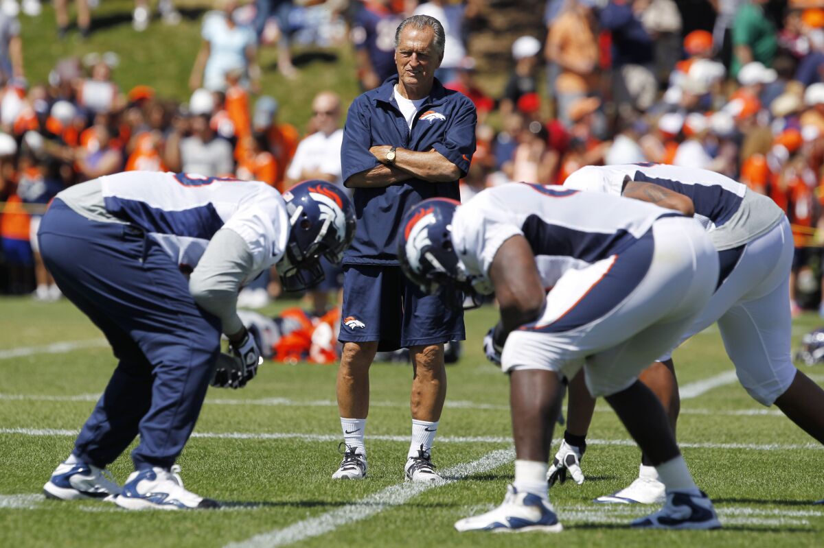FILE - Denver Broncos offensive line consultant Alex Gibbs, back, looks on as linemen take part in drills after the morning session at the team's NFL training camp in Englewood, Colo., in this Tuesday, Aug. 6, 2013, file photo. Gibbs, the innovative offensive line coach whose zone-blocking scheme helped lead the Denver Broncos to back-to-back Super Bowl triumphs in the 1990s, has died at age 80, the team said. The team said Gibbs died Monday, July 12, 2021, from complications of a stroke with family by his side in his Phoenix home. (AP Photo/David Zalubowski, File)