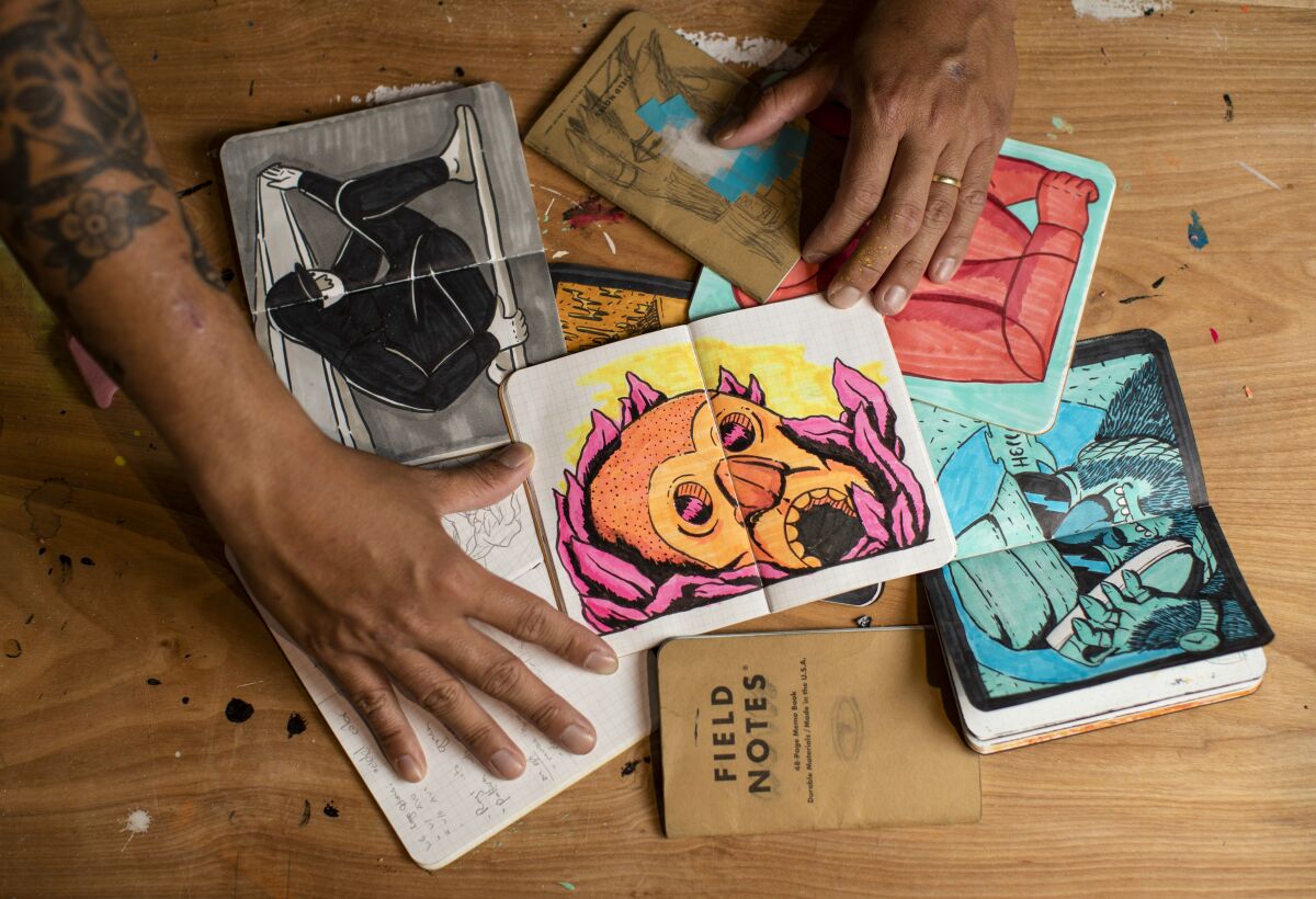 A man's hand and tattooed arm with notebooks opened to colorful sketches.