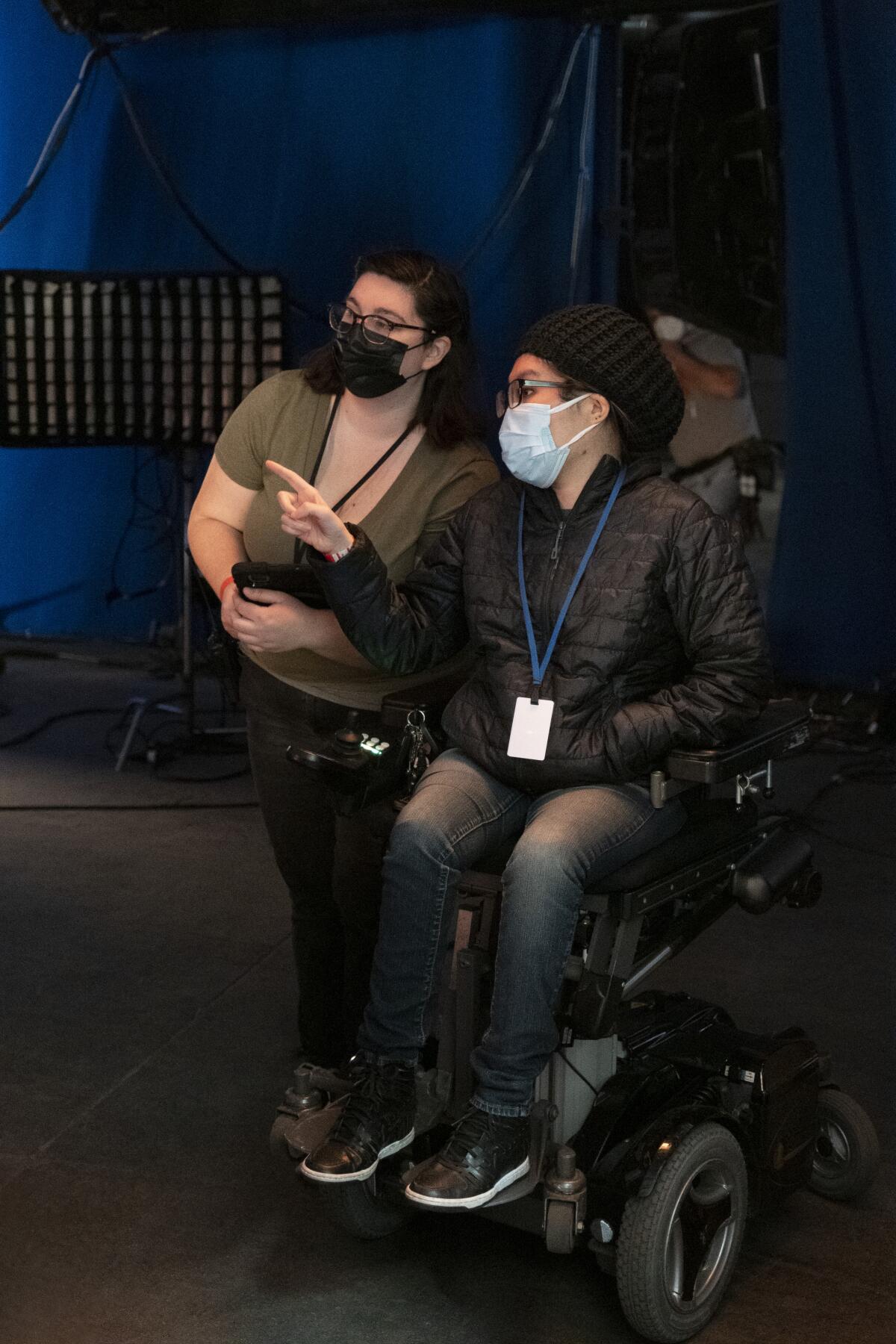 Two masked women, one sitting and one standing, talk on a film set.