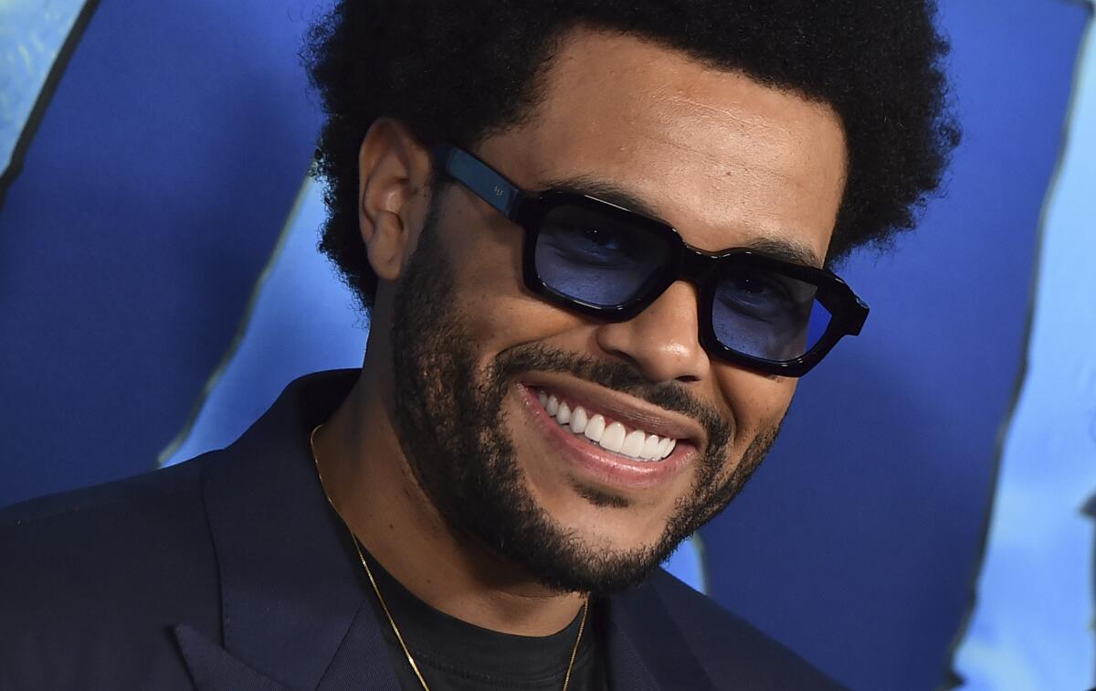 A man wears dark sunglasses and a dark suit as he smiles against a blue backdrop