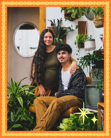 A woman perches on the arm of a chair in which is seated her brother. Her arm is around his shoulders. Plants surround them.