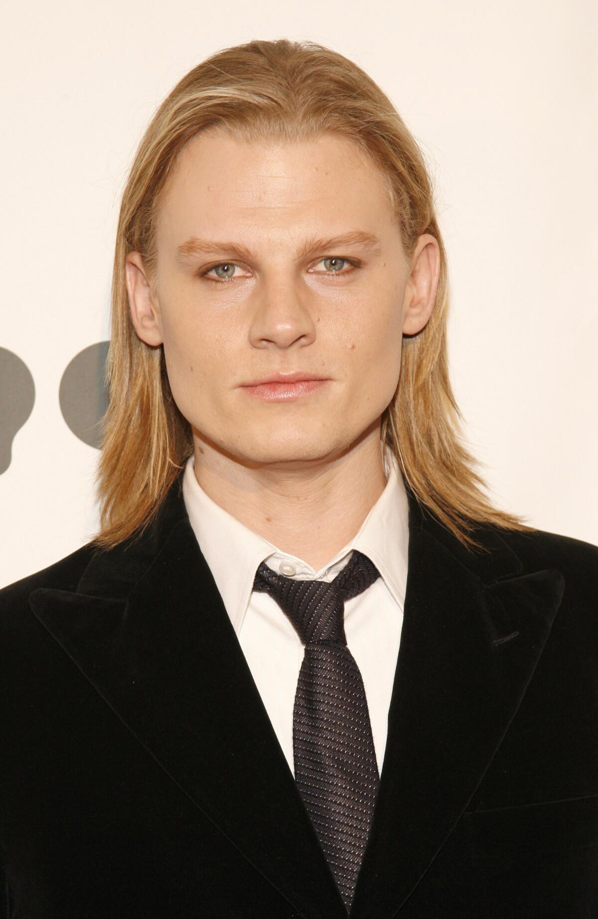 Jeffrey Carlson, with shoulder-length blond hair, wears a black velvet blazer, white shirt and tie. 