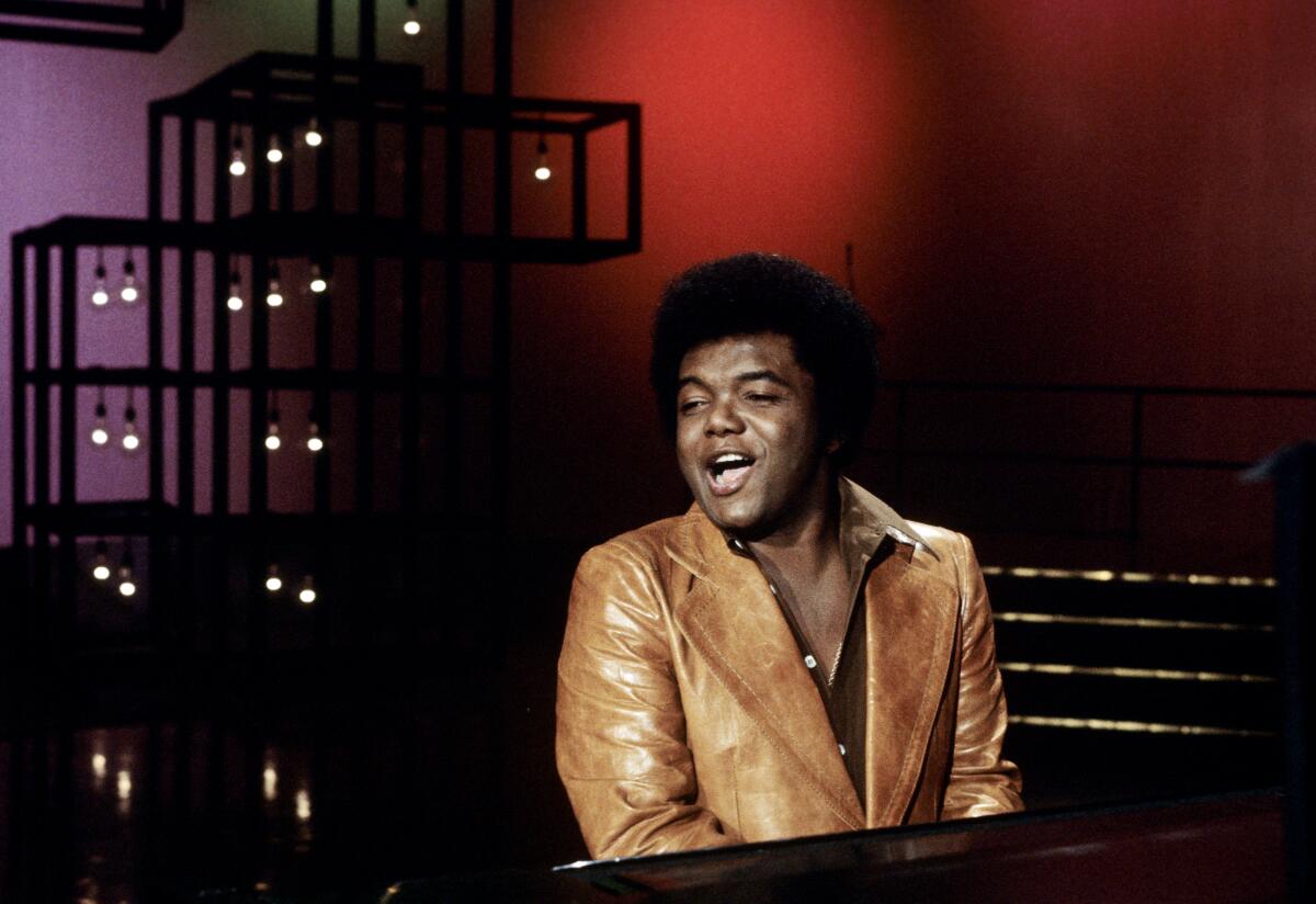 A man in a light brown leather jacket plays the piano and sings 