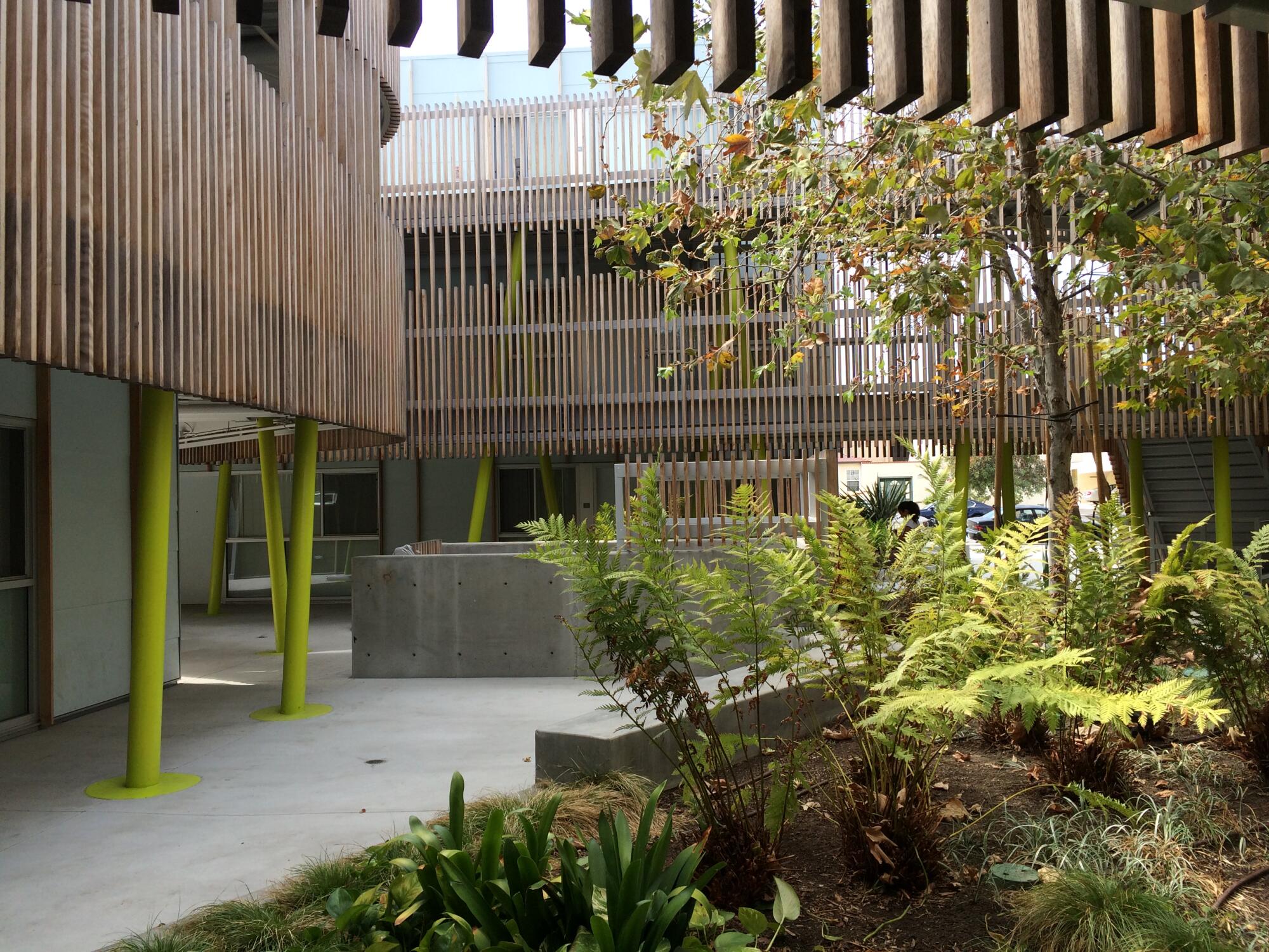Trees and ferns are seen in the courtyard of a two-story building whose balconies are lined with wood-slat balustrades.