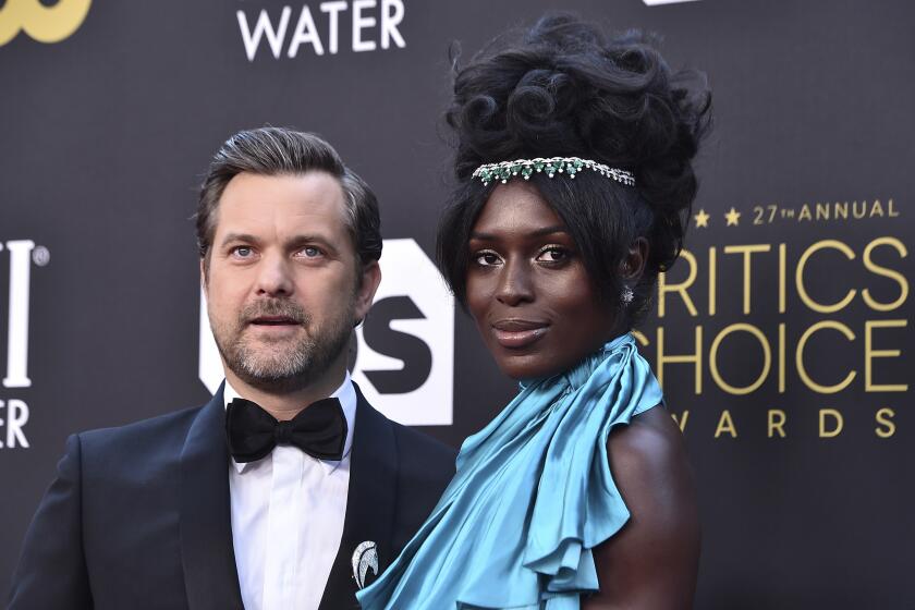 Joshua Jackson poses in a black tuxedo and bowtie next to Jodie Turner-Smith posing in a blue dress and matching headband.